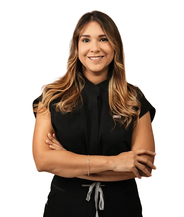 Una mujer joven con cabello castaño largo y reflejos rubios sonríe a la cámara, vestida con un uniforme médico negro y los brazos cruzados, sobre un fondo color canela.