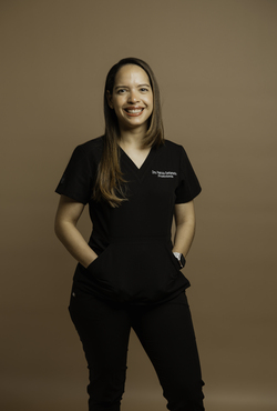 Una mujer joven con cabello castaño largo sonríe a la cámara, vestida con un uniforme médico negro, sobre un fondo color canela.