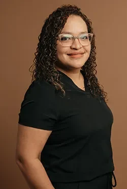 Mujer con cabello rizado y gafas, sonriendo, vestida con un uniforme médico negro, con una mano en el bolsillo, de pie contra un fondo marrón.