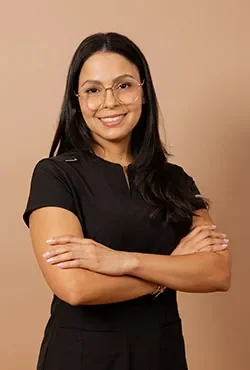 Mujer sonriente con cabello oscuro y gafas, vestida con uniforme médico negro, posa con los brazos cruzados sobre un fondo beige.